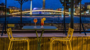 Une table avec deux chaises et deux boissons, placée sur une terrasse en bois donnant sur une rivière, avec un pont et des bâtiments en arrière-plan.