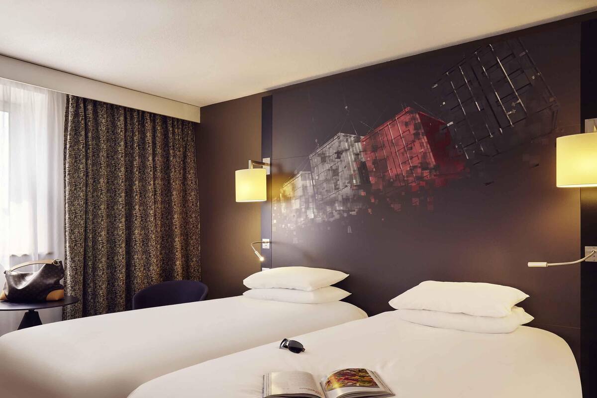 Une chambre d’hôtel avec deux lits, une grande fenêtre avec des rideaux, une lampe fixée au mur et un élément décoratif mural.