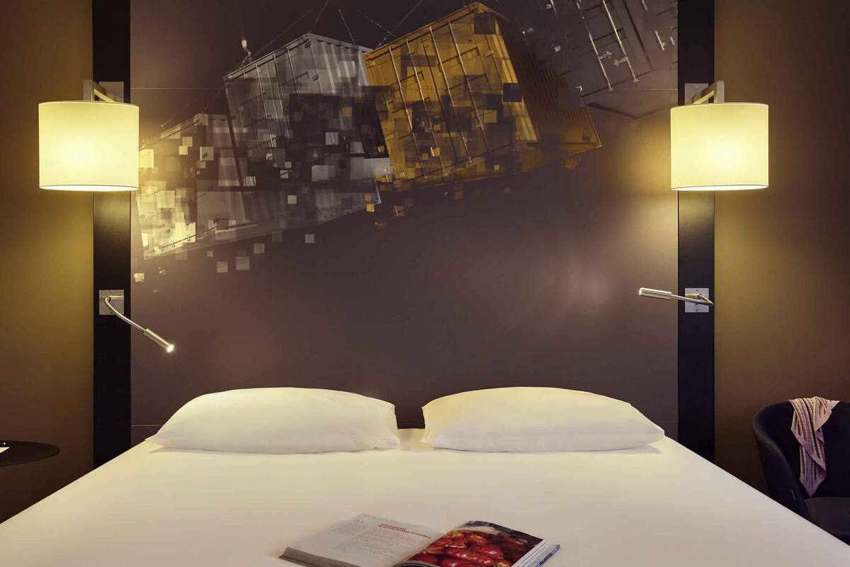 Une chambre d’hôtel avec un grand lit, deux lampes de chevet, un téléviseur fixé au mur et un magazine posé sur le lit.
