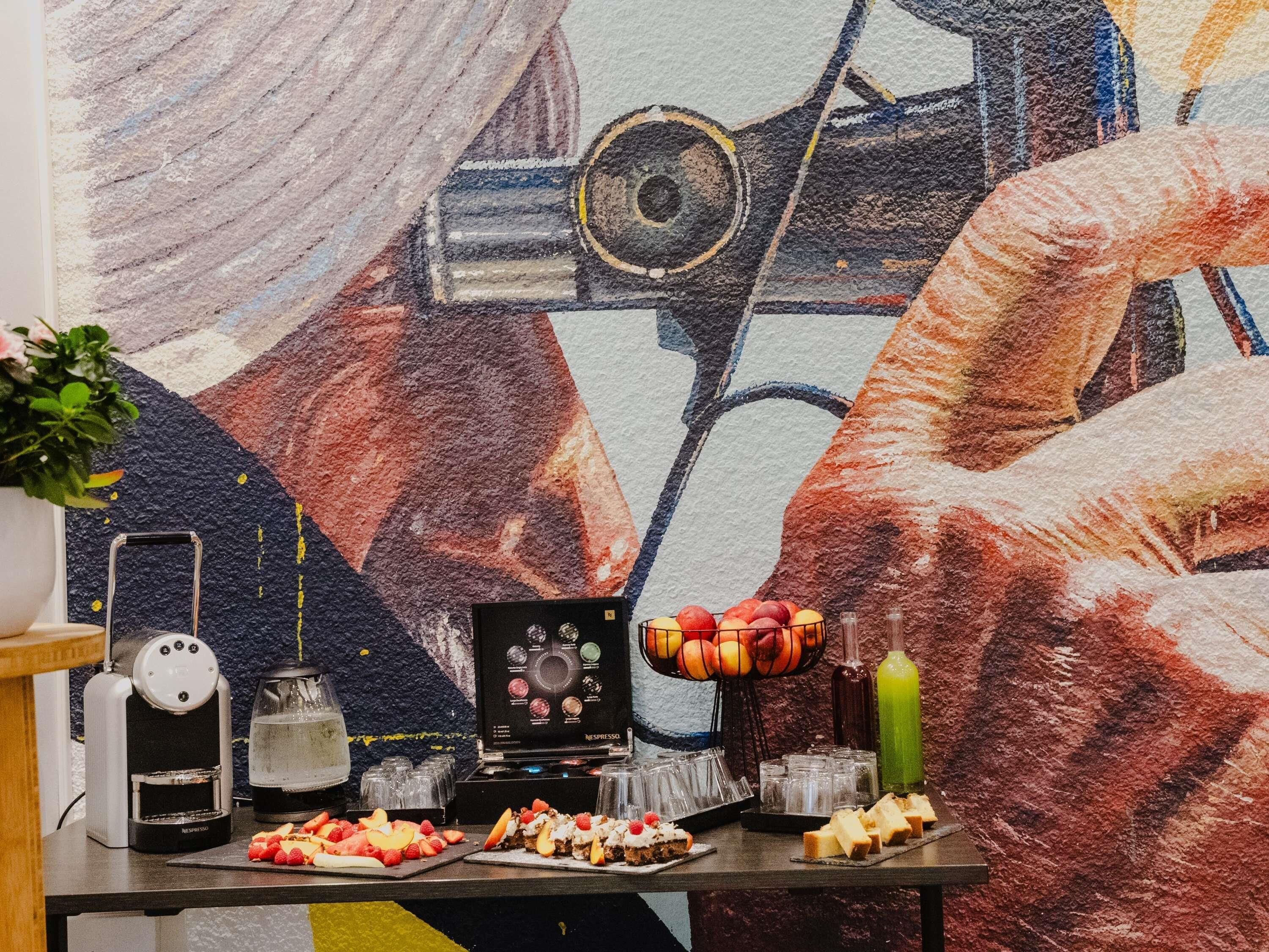 Taula amb una cafetera, una màquina de Nespresso, una cistella de fruita, un got de suc i diferents aperitius.