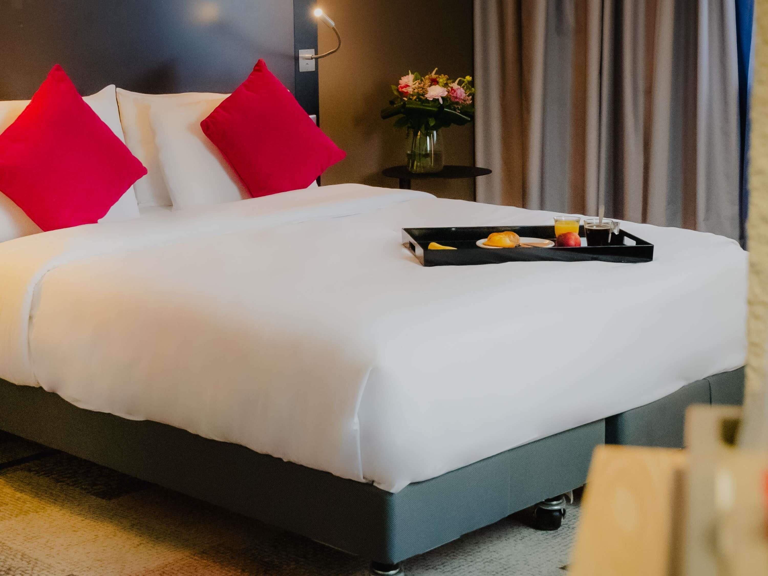 Una cama bien hecha con ropa blanca y almohadas rojas, una bandeja con alimentos para el desayuno y un florero con flores sobre una mesita de noche.