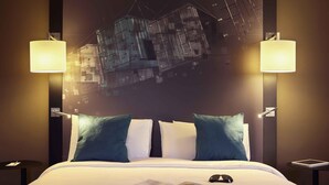 Une chambre d’hôtel avec un grand lit, deux lampes fixées au mur, une table de chevet, une tablette et un livre.