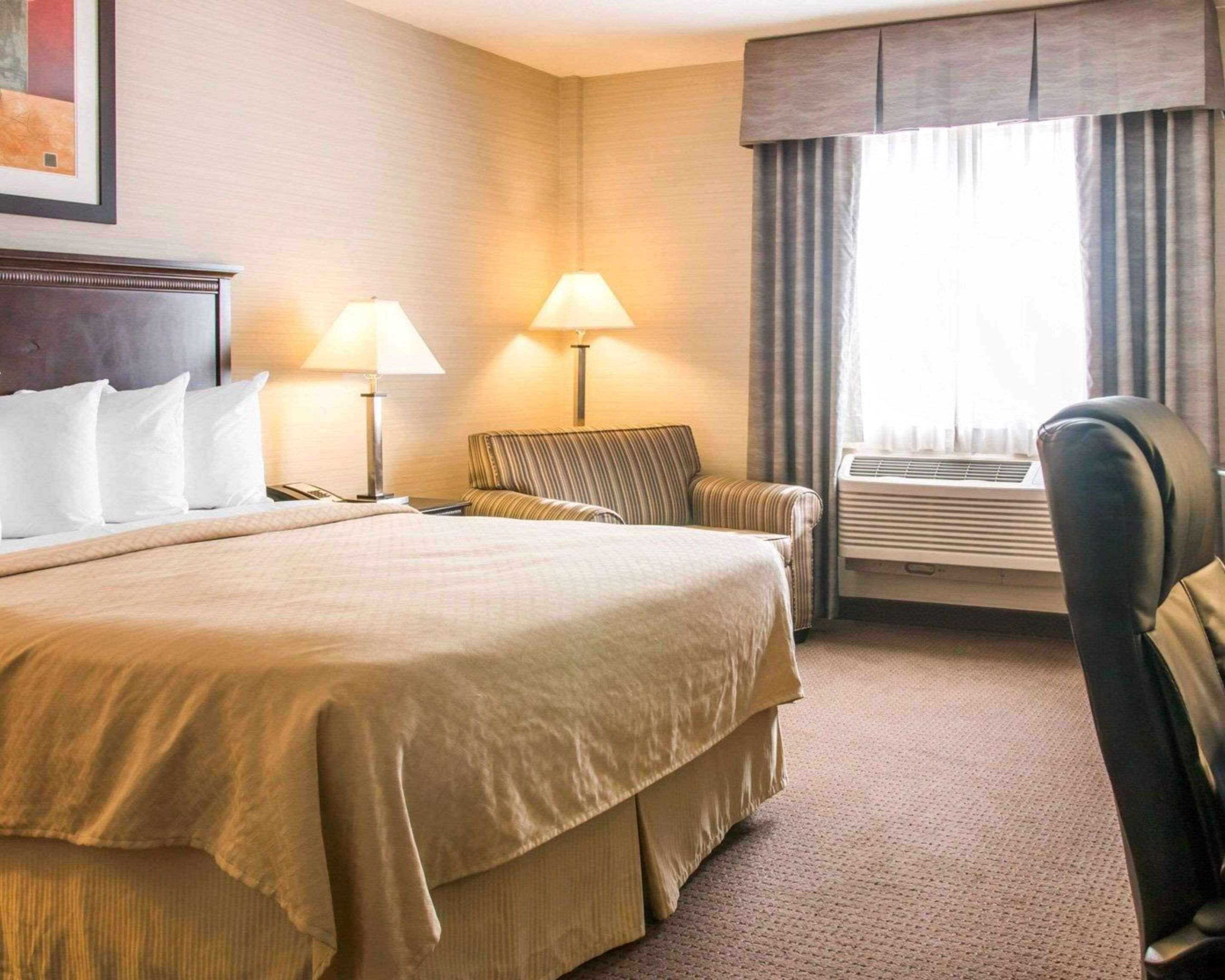 A hotel room with a large bed, two bedside lamps, a chair, an air conditioning unit, and a window with curtains.