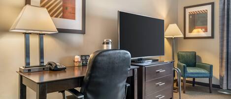 Habitación de hotel con escritorio, una silla, televisor, lámpara, cafetera e imagen enmarcadas en la pared.