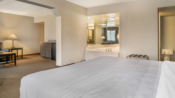 Habitación de hotel con una cama grande, un escritorio con lámpara, una kitchenette y un baño con espejo y lavamanos.