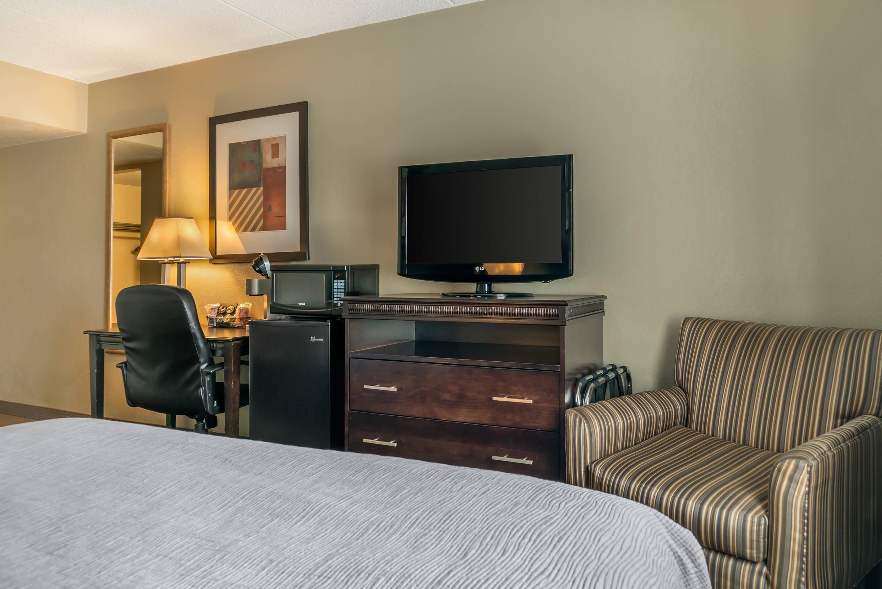 A hotel room with a bed, a desk with a computer and a television, a striped armchair, and a lamp.
