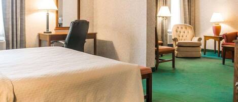 A hotel room with a large bed, a desk with a chair, a separate armchair, and a small table with a lamp.