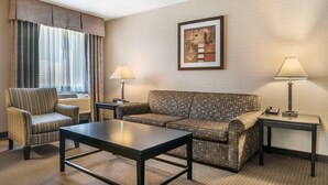 Habitación de hotel con sofá, sillón y una mesa. Hay una lámpara en una mesita auxiliar y un cuadro en la pared.