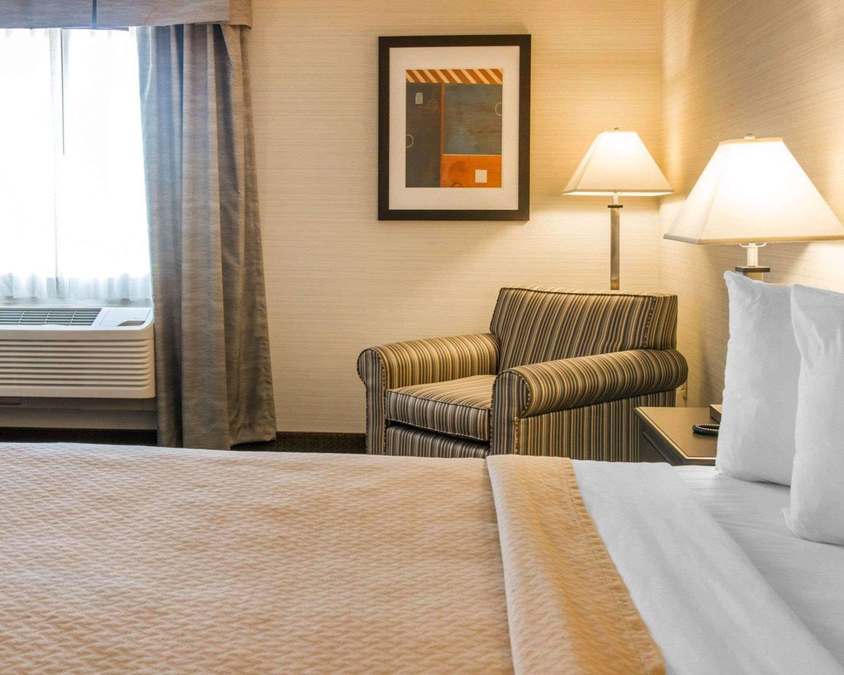 Een hotelkamer met een bed, een gestreept fauteuil, een nachtkastje met een lamp, en een lijst met een afbeelding aan de muur.