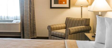 Een hotelkamer met een bed, een gestreept fauteuil, een nachtkastje met een lamp, en een lijst met een afbeelding aan de muur.