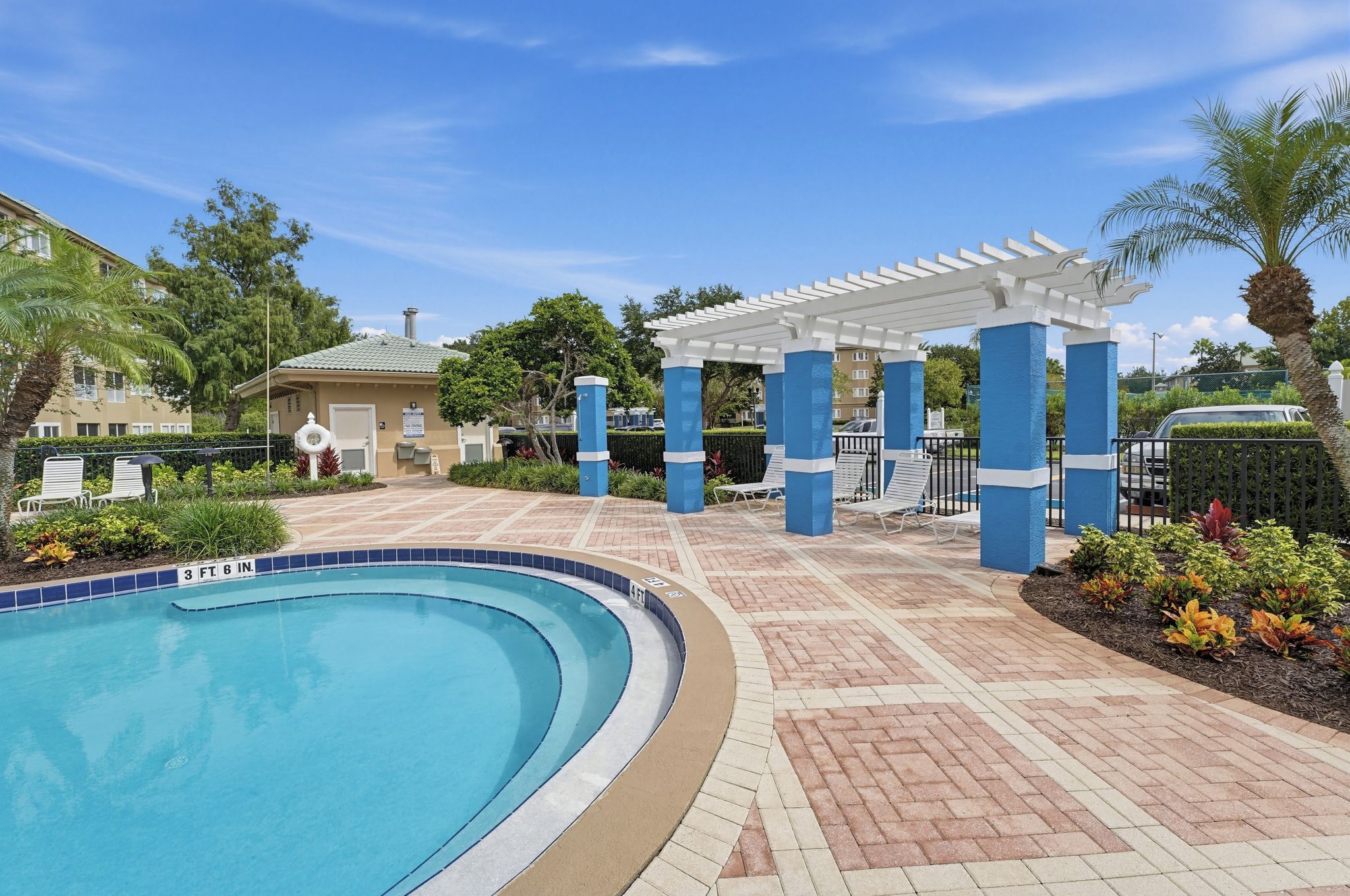 Une piscine bordée de bleu, un patio couvert avec des colonnes blanches, et un bâtiment en arrière-plan.