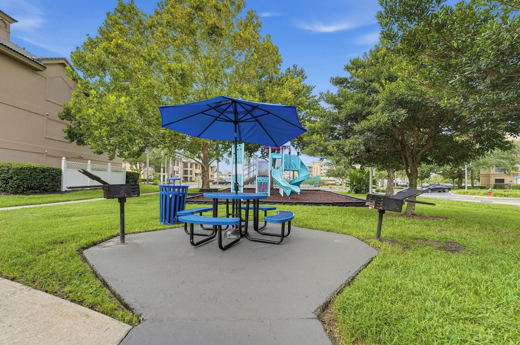 An outdoor seating area with a table and chairs, a blue umbrella, and a playground in the background.