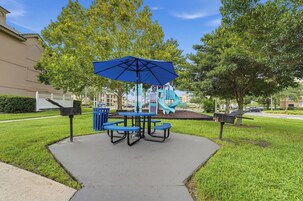 Un área de estar al aire libre con una mesa, sillas, una sombrilla azul y un parque infantil al fondo.