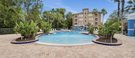 A swimming pool with lounge chairs and palm trees, surrounded by a brick walkway and a building with balconies.