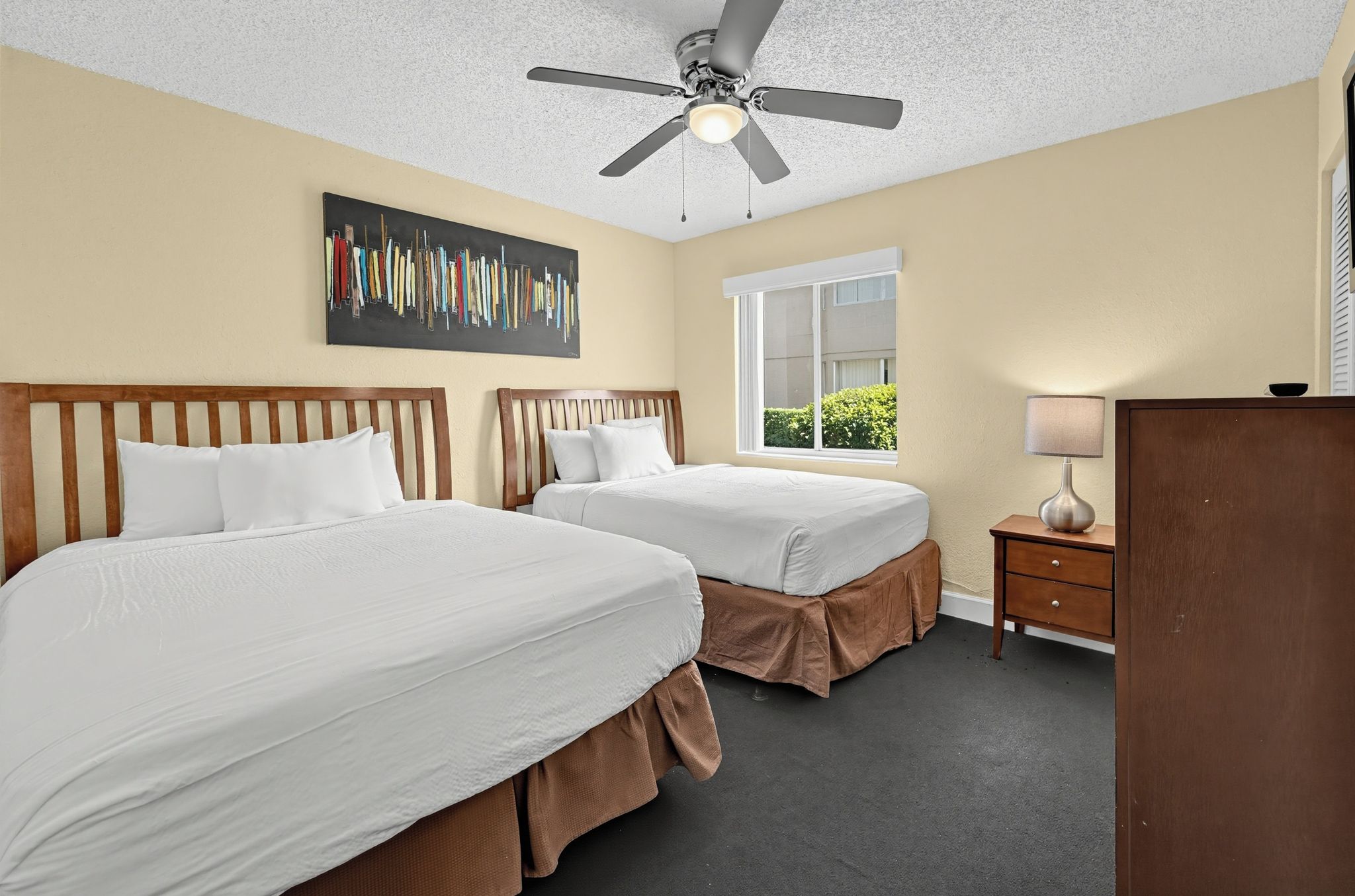 A hotel room with two beds, a ceiling fan, a window with a view, and a desk with a lamp.
