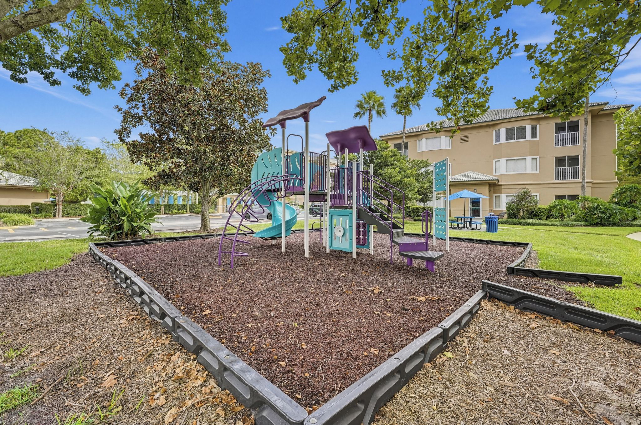 A playground with a slide and climbing structures, surrounded by mulch and trees.