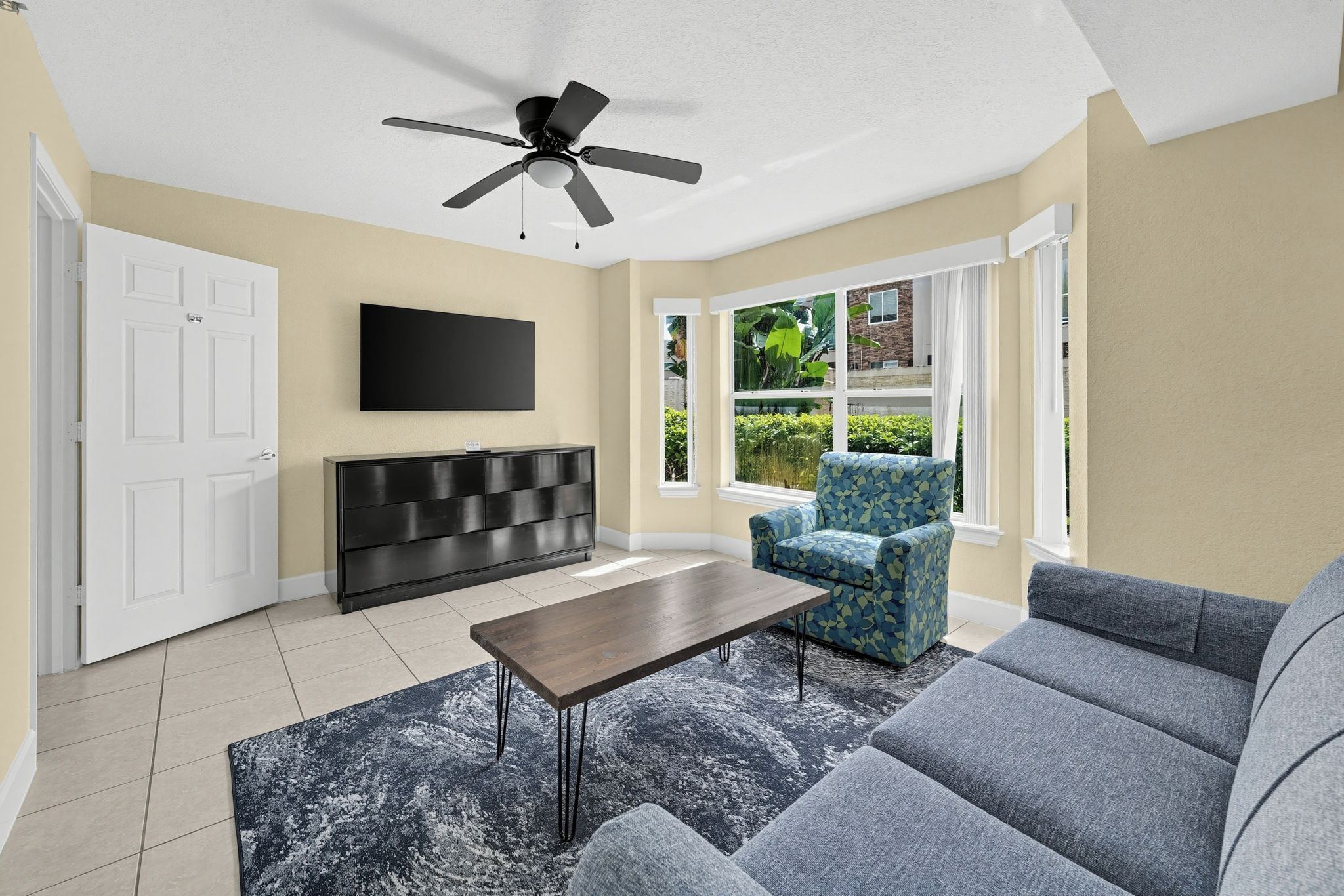 A living room with a ceiling fan, a flat-screen TV mounted on the wall, a black entertainment unit, a coffee table, a patterned armchair, and a grey sofa.