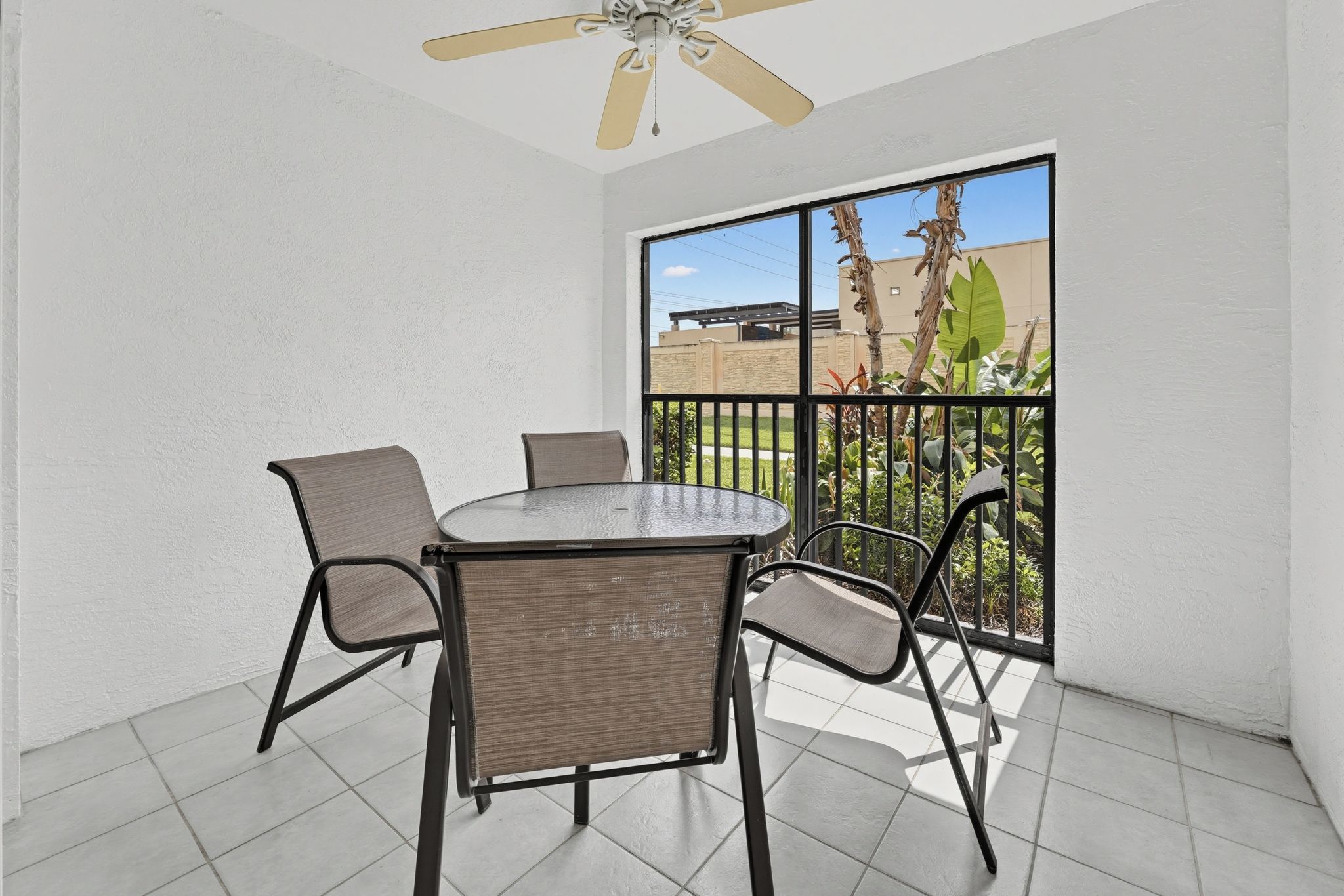 A balcony with a round table and chairs, a ceiling fan, and a view of a building and greenery.