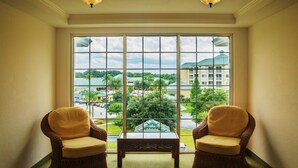 A room with two wicker chairs and a small table, a large window with a view of a building and greenery, and two wall-mounted lights.