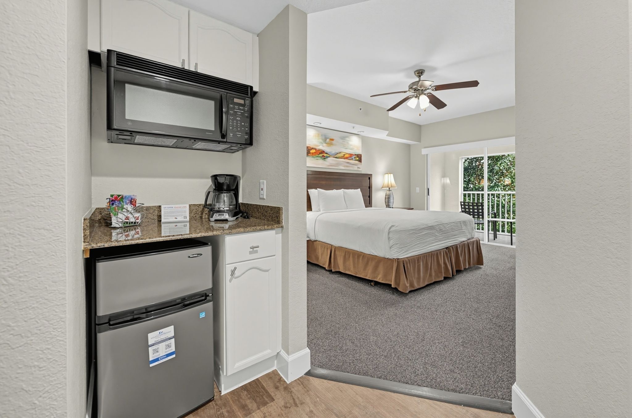 A compact hotel room with a kitchenette, a bed with a brown skirt, a ceiling fan, and a view through a glass door.