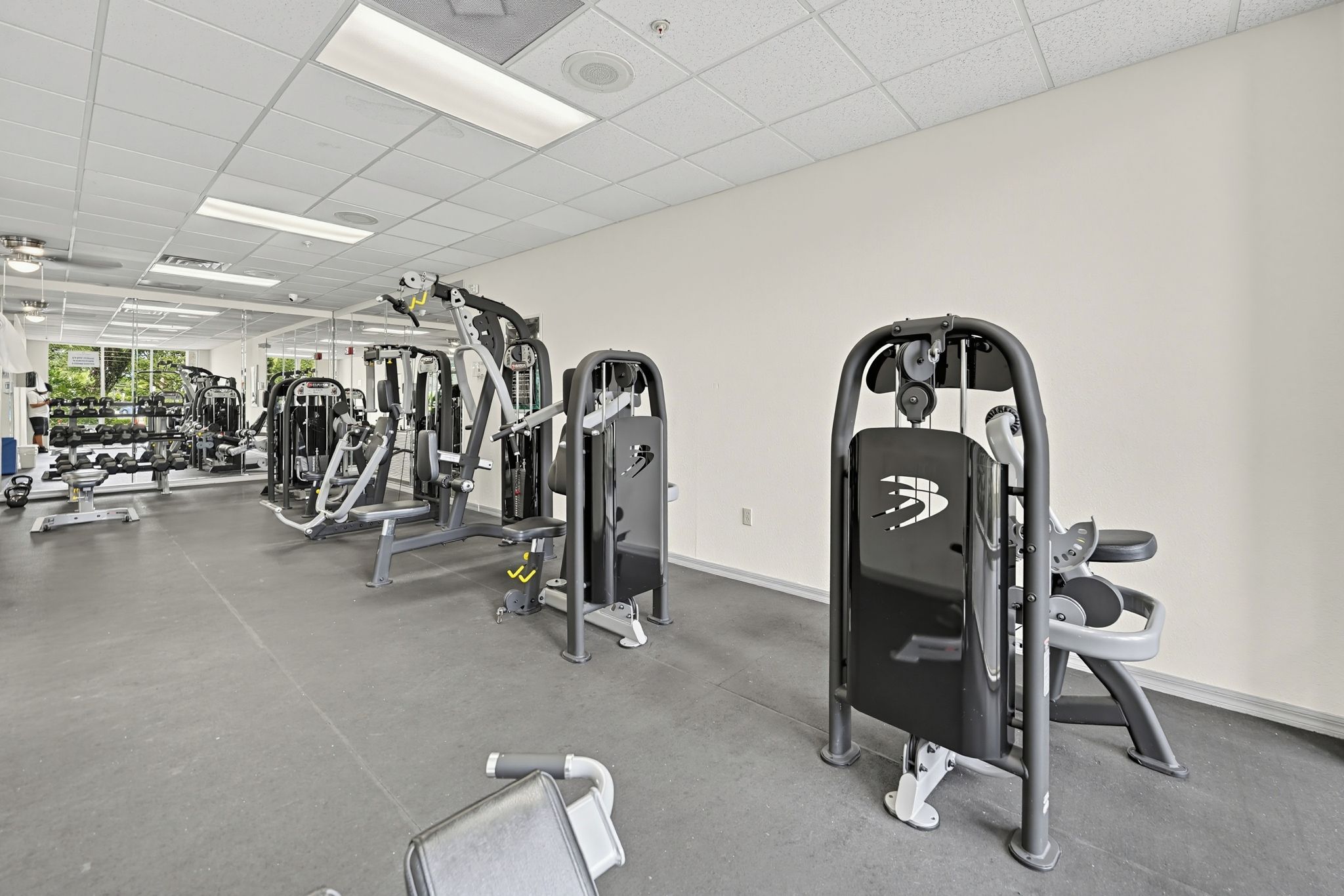 Ein Fitnessstudio mit verschiedenen Trainingsgeräten, einer Decke mit eingelassener Beleuchtung und Blick ins Freie durch große Fenster.