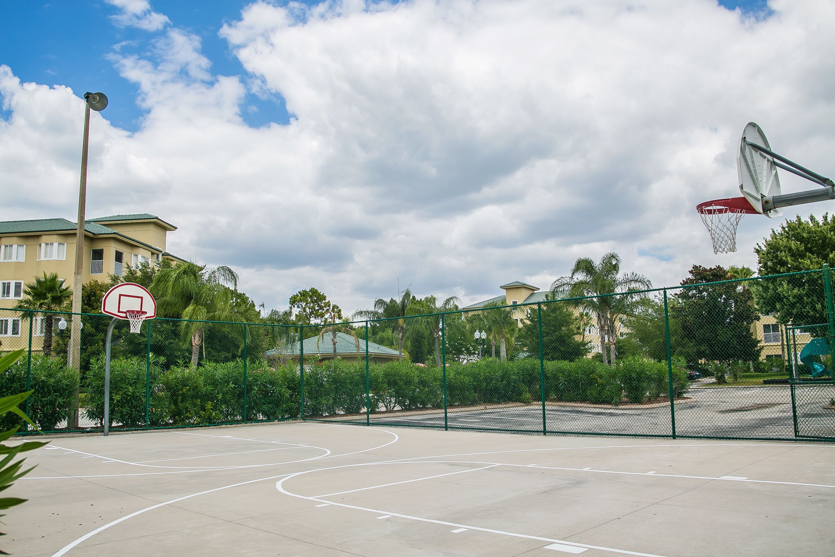 An outdoor basketball court with a hoop, surrounded by a fence and greenery.