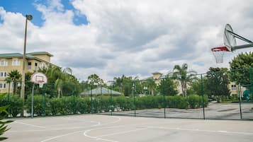 An outdoor basketball court with a hoop, surrounded by a fence and greenery.