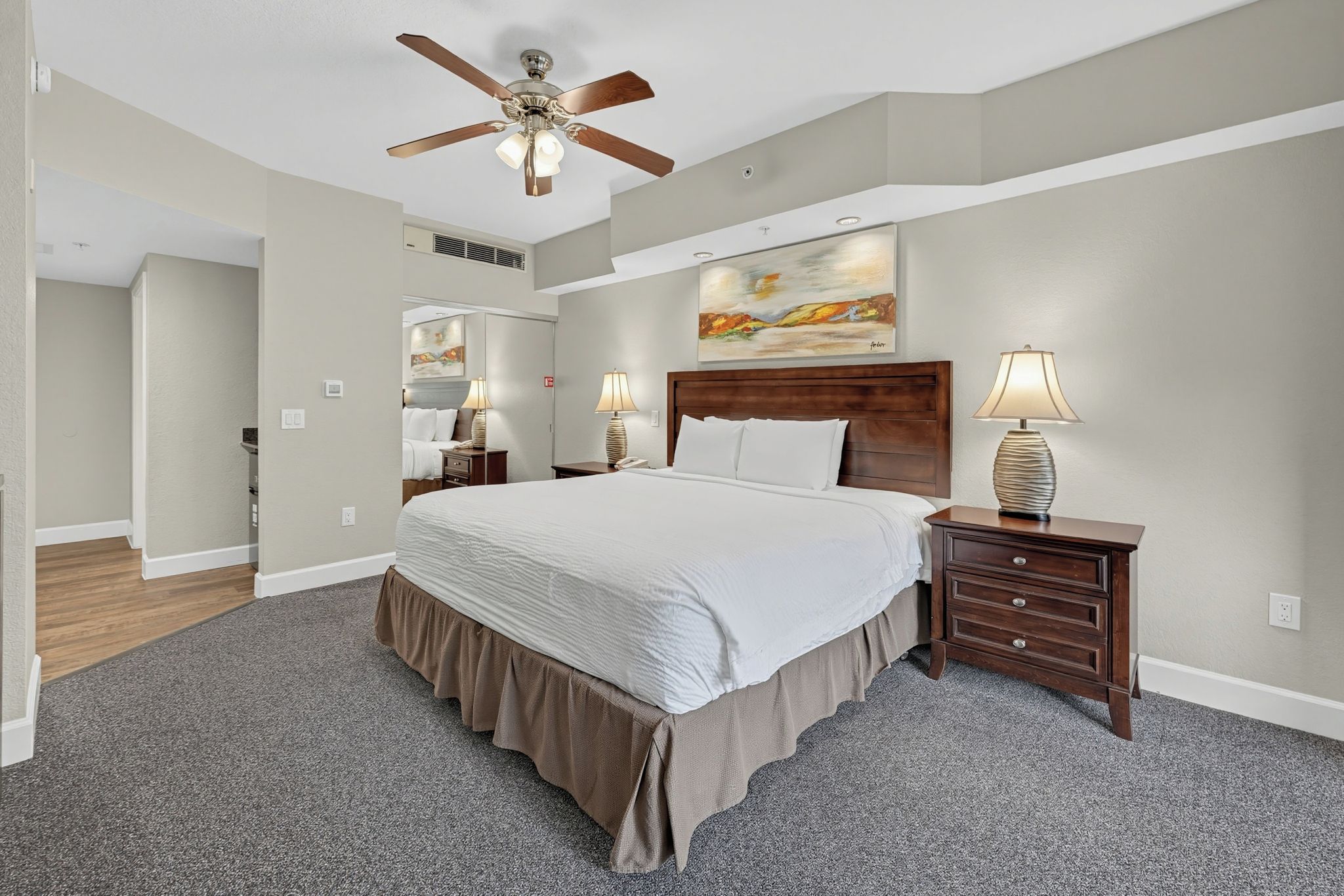 A hotel room with a large bed, two bedside tables with lamps, a ceiling fan, and a painting on the wall.