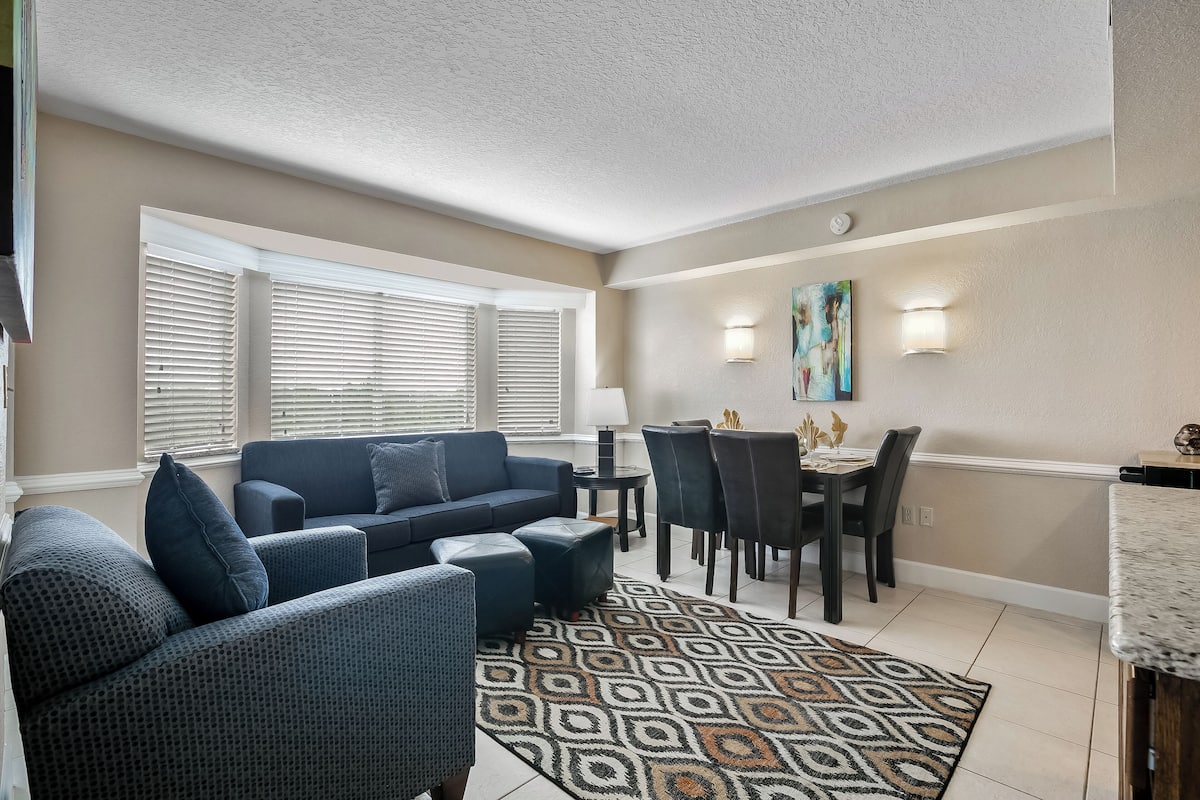 A living room with a blue sofa, ottoman, and chairs, a patterned rug, a dining area with a table and chairs, and a painting on the wall.