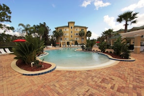 A pool area with lounge chairs and umbrellas, surrounded by palm trees and a building with a green roof.