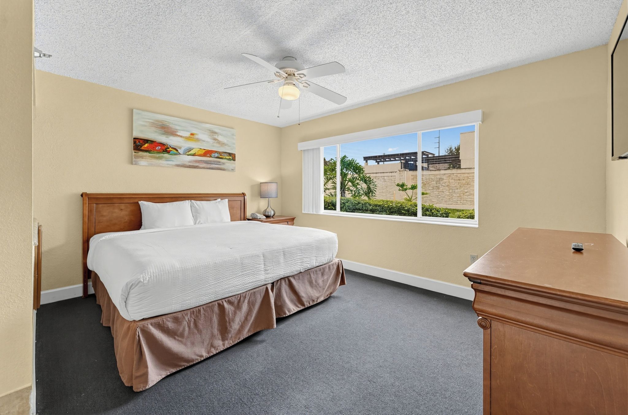 Une chambre d’hôtel avec un lit, une table de chevet, un bureau, un ventilateur de plafond, une fenêtre donnant sur un paysage agréable et un tableau au mur.
