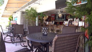 Un'area esterna con posti a sedere, sedie in vimini e un tavolo rotondo, un bar con diversi articoli e uno schermo televisivo.