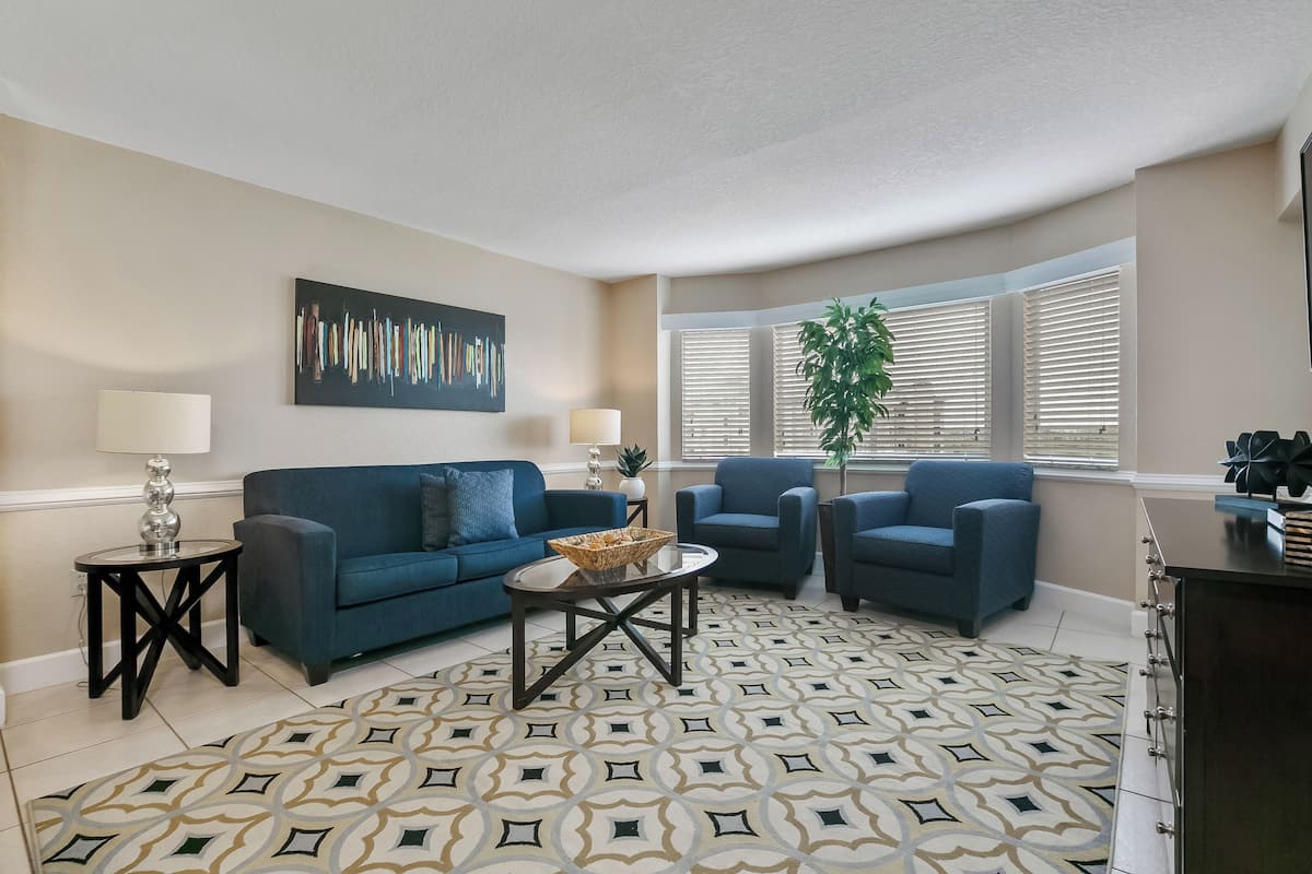A living room with a blue sofa, two armchairs, a coffee table, a patterned rug, and a large abstract painting on the wall.