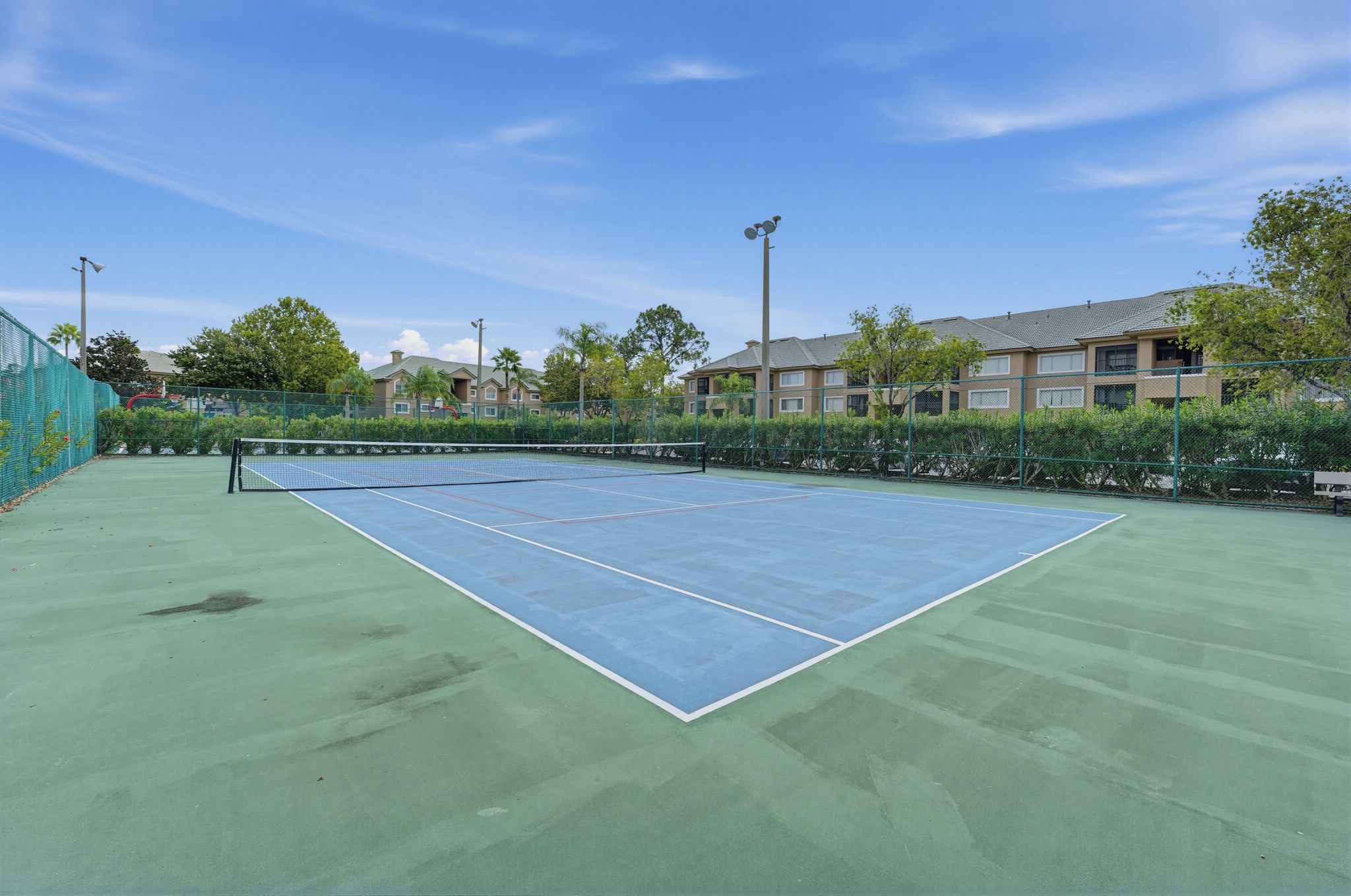 An outdoor tennis court with a net, surrounded by greenery and buildings.