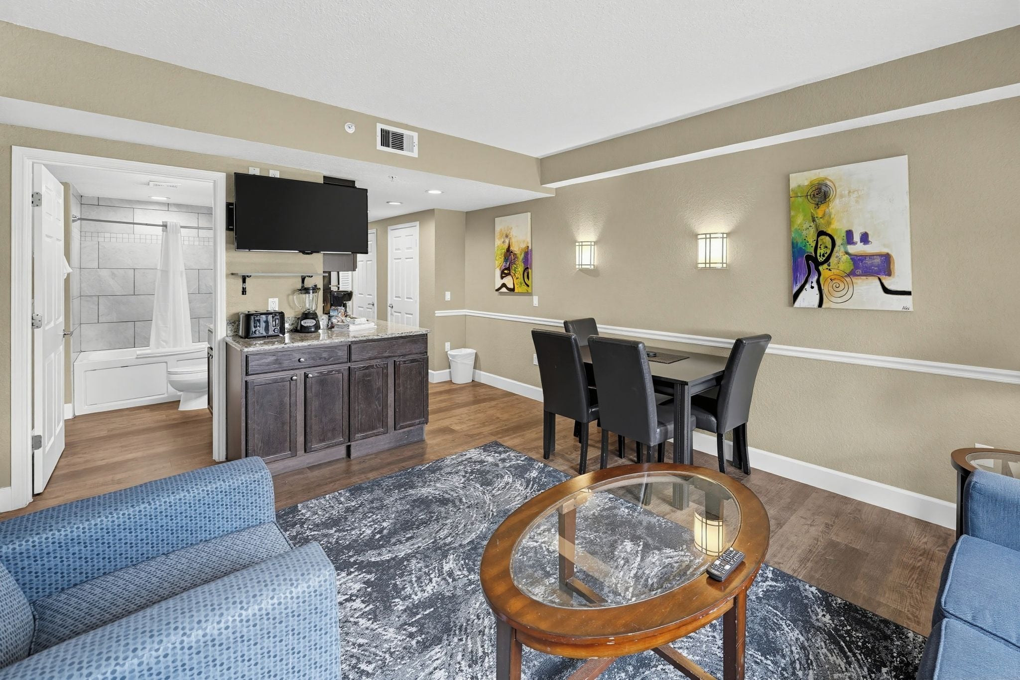 A modern living room with a dining area, a kitchen with a flat-screen TV, and artwork on the walls.