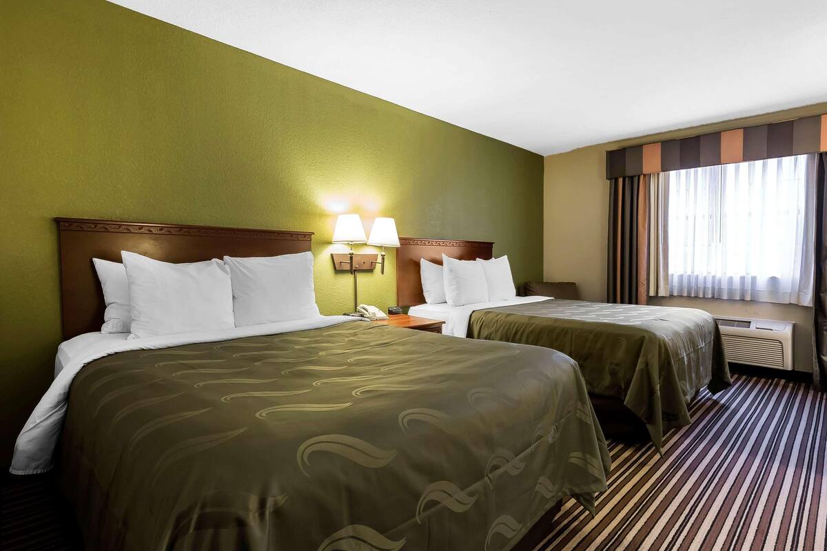 Ein Hotelzimmer mit zwei Betten, einem Nachttisch mit Lampen, einem Telefon und einem Fenster mit Vorhängen.
