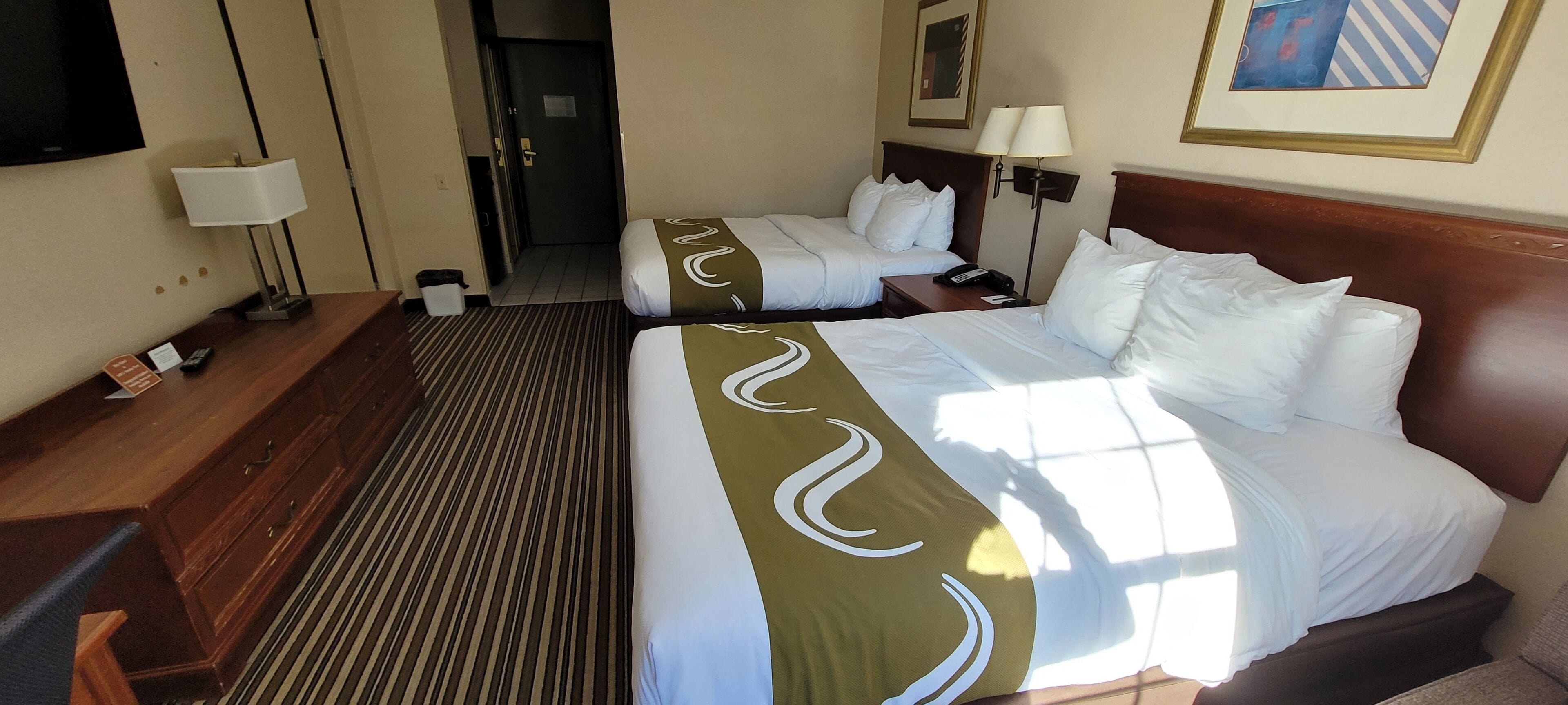 A hotel room with two beds, a desk, a chair, a lamp, and a television.
