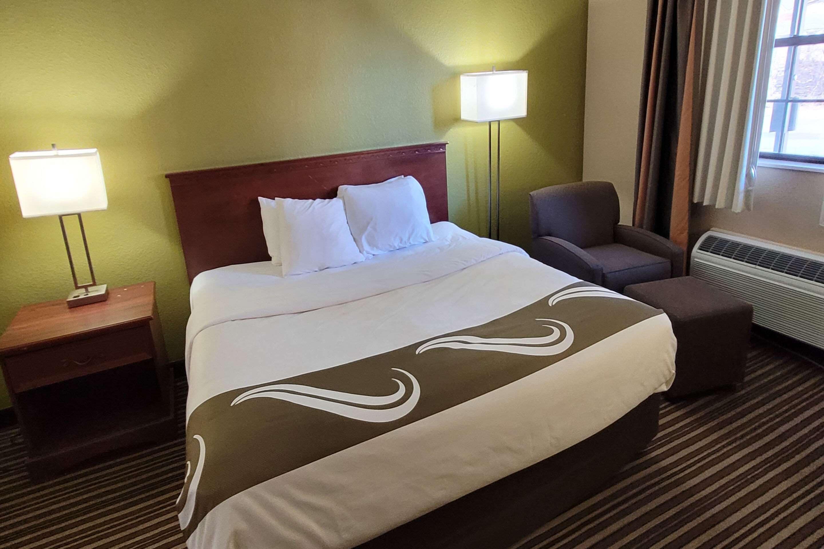 A hotel room with a large bed, two bedside lamps, a chair, and a heater.