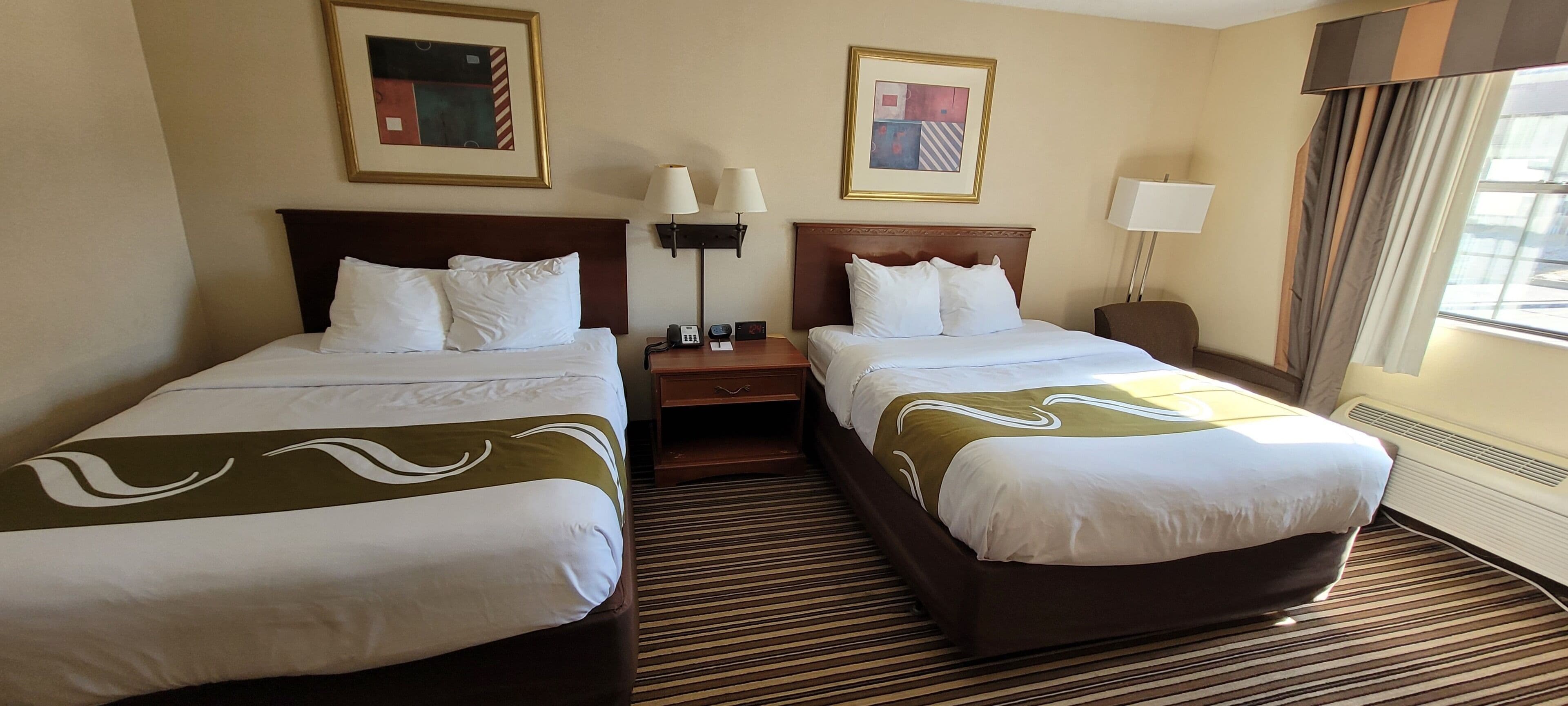 A hotel room with two beds, a nightstand, a lamp, and two framed pictures on the wall.
