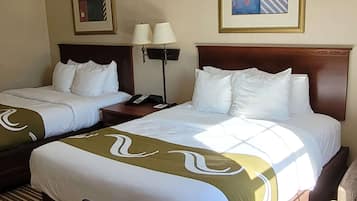 A hotel room with two beds, each with white bedding and a green decorative runner. There are two bedside tables, one with a lamp and the other with a phone. The room has a wooden headboard and a framed picture on the wall.