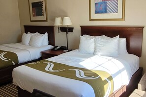 A hotel room with two beds, each with white bedding and a green decorative runner. There are two bedside tables, one with a lamp and the other with a phone. The room has a wooden headboard and a framed picture on the wall.