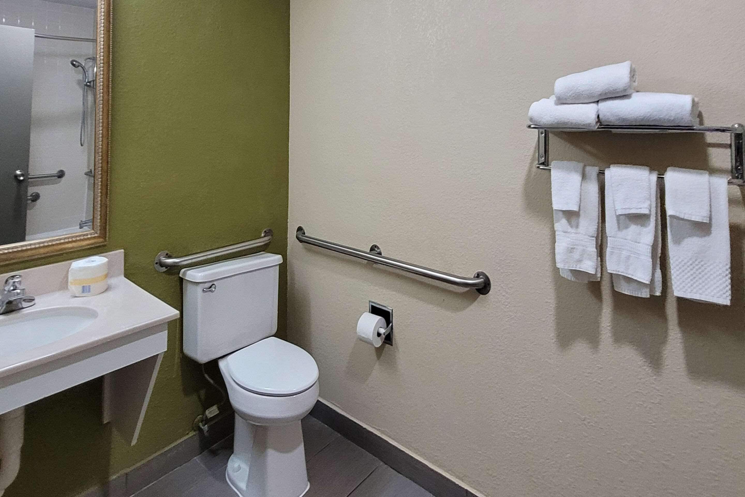 A bathroom with a toilet, sink, mirror, towel rack, and handrails.