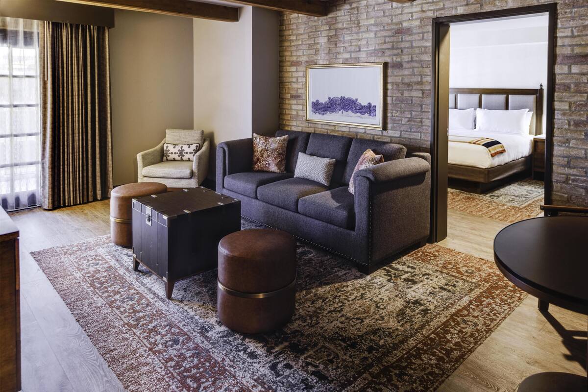 A modern hotel room with a grey sofa, a small round table, a wooden ottoman, a large rug, and a bed with white linens.