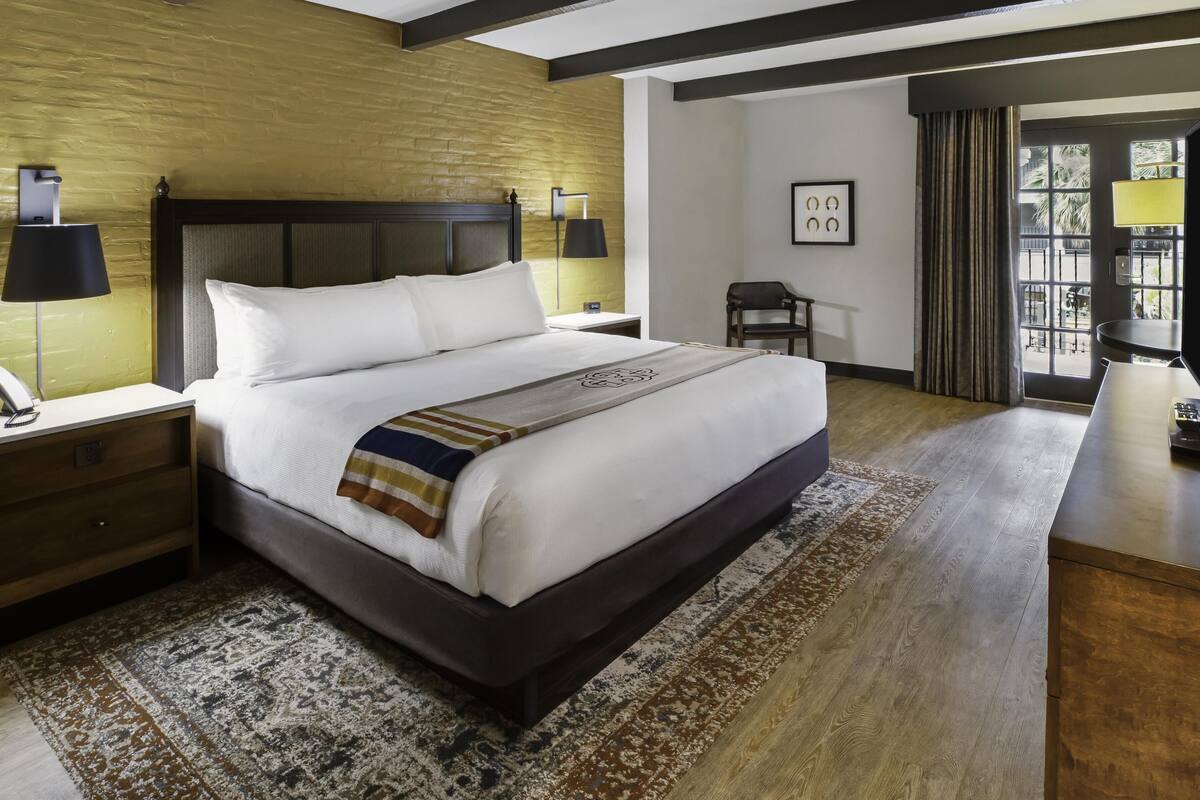 A hotel room with a large bed, a bedside table with a lamp, a nightstand, a chair, a desk, and a window with curtains.