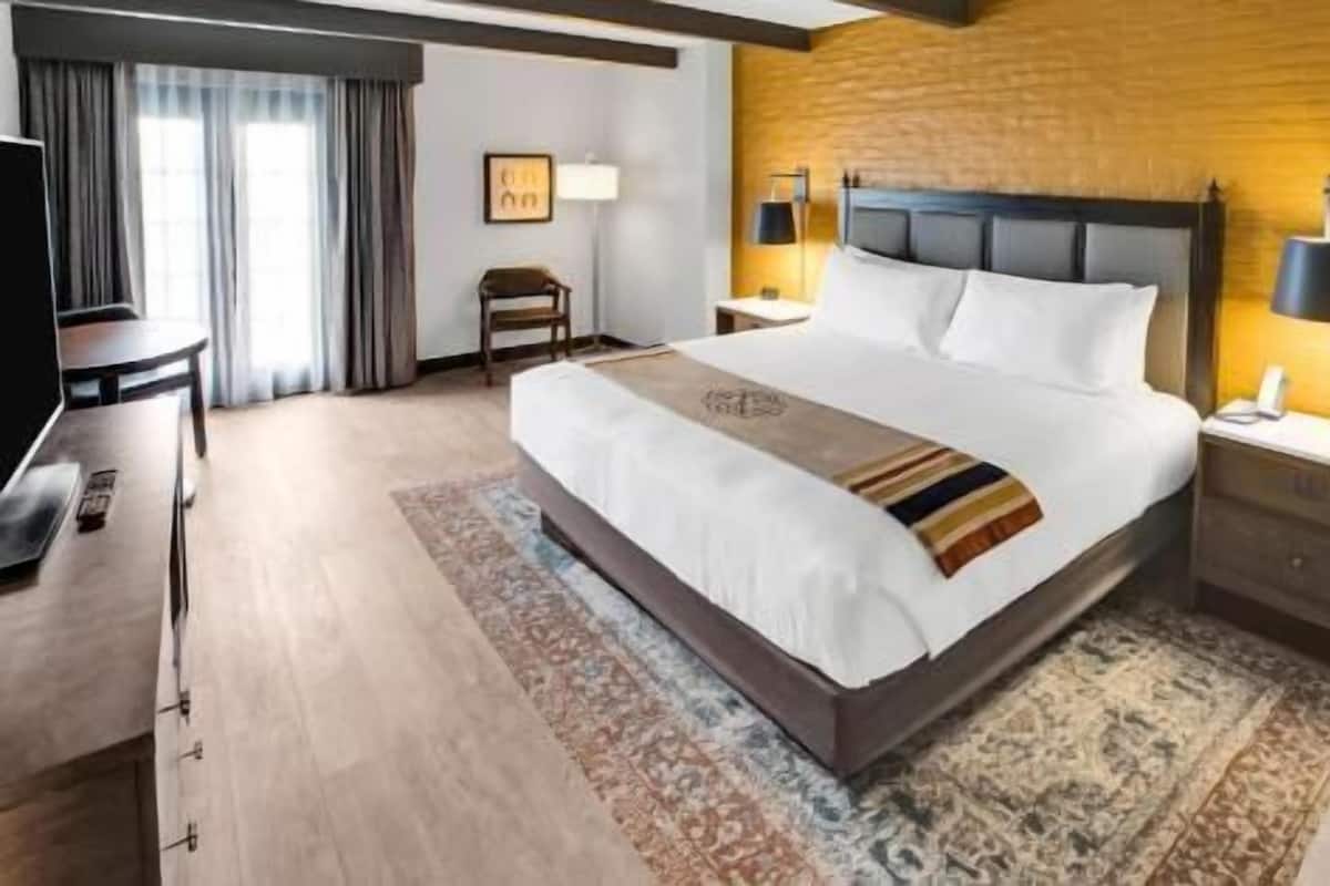 A hotel room with a large bed, a desk, a chair, a TV, and a lamp.