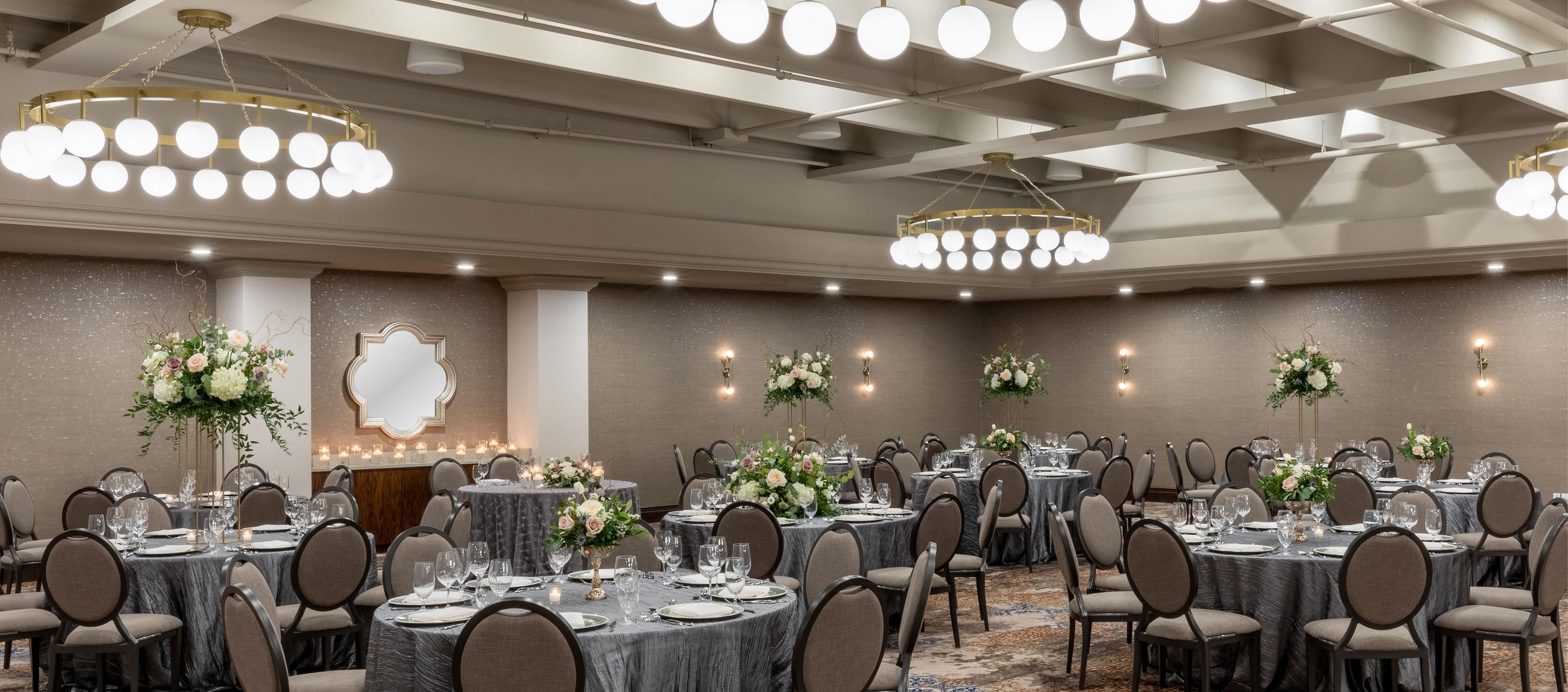 Ein Festsaal mit runden Tischen, Stühlen, einer Kristalllüster und Blumendekorationen sowie Wandleuchten.