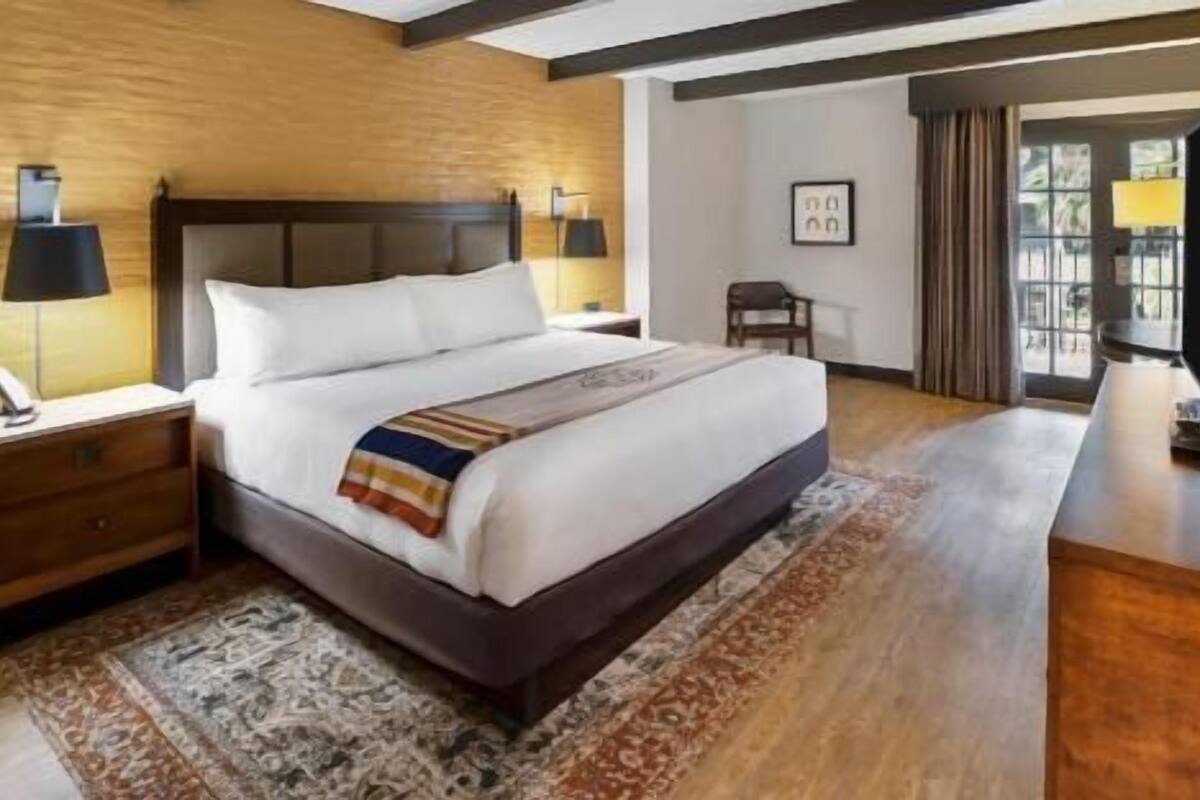 A hotel room with a large bed, wooden headboard, two bedside tables with lamps, a small desk, and a large window with curtains.
