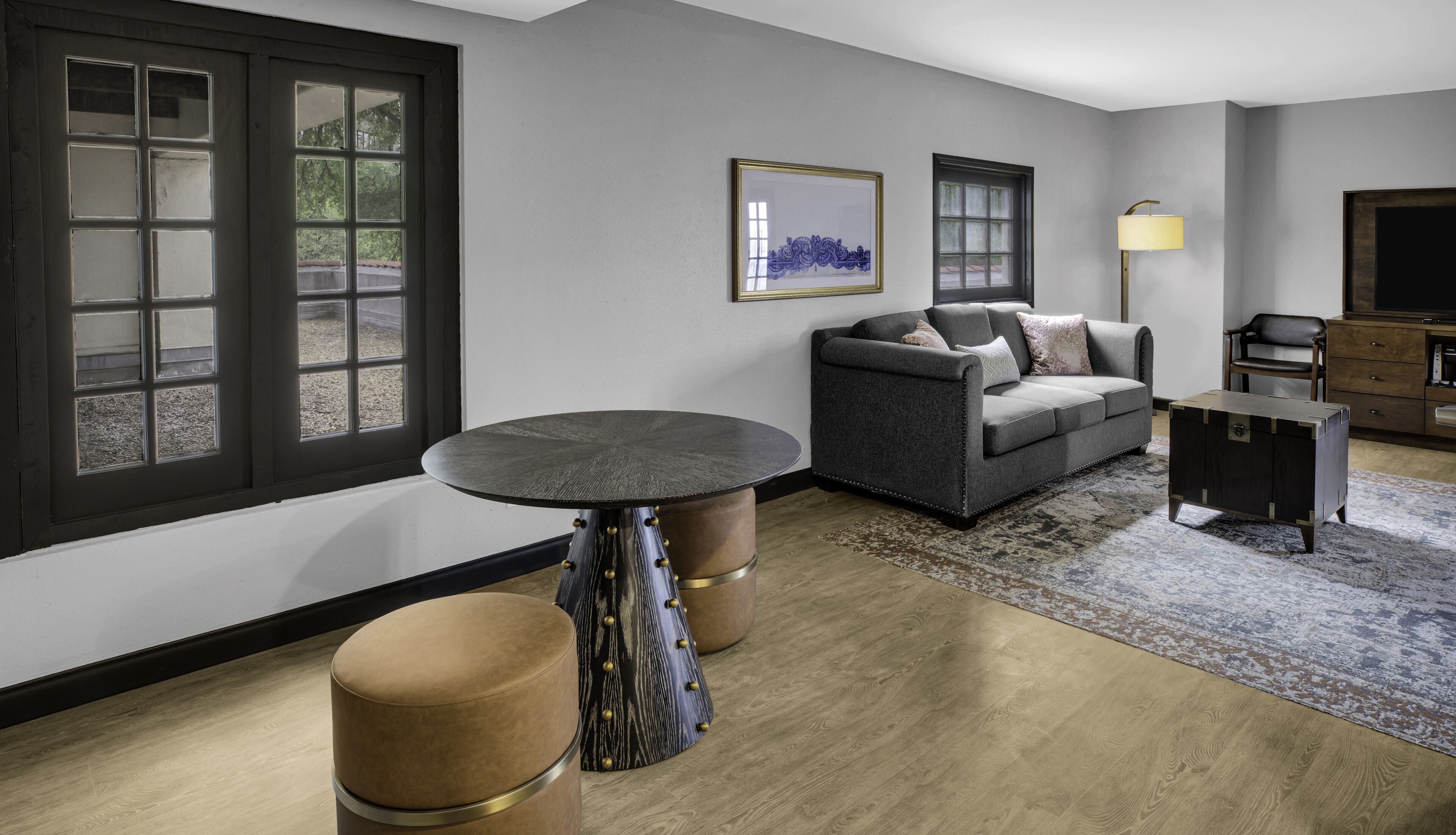 A modern living room with a grey sofa, a round table, and a coffee table. There is a window with a view of trees, a framed artwork on the wall, and a lamp.