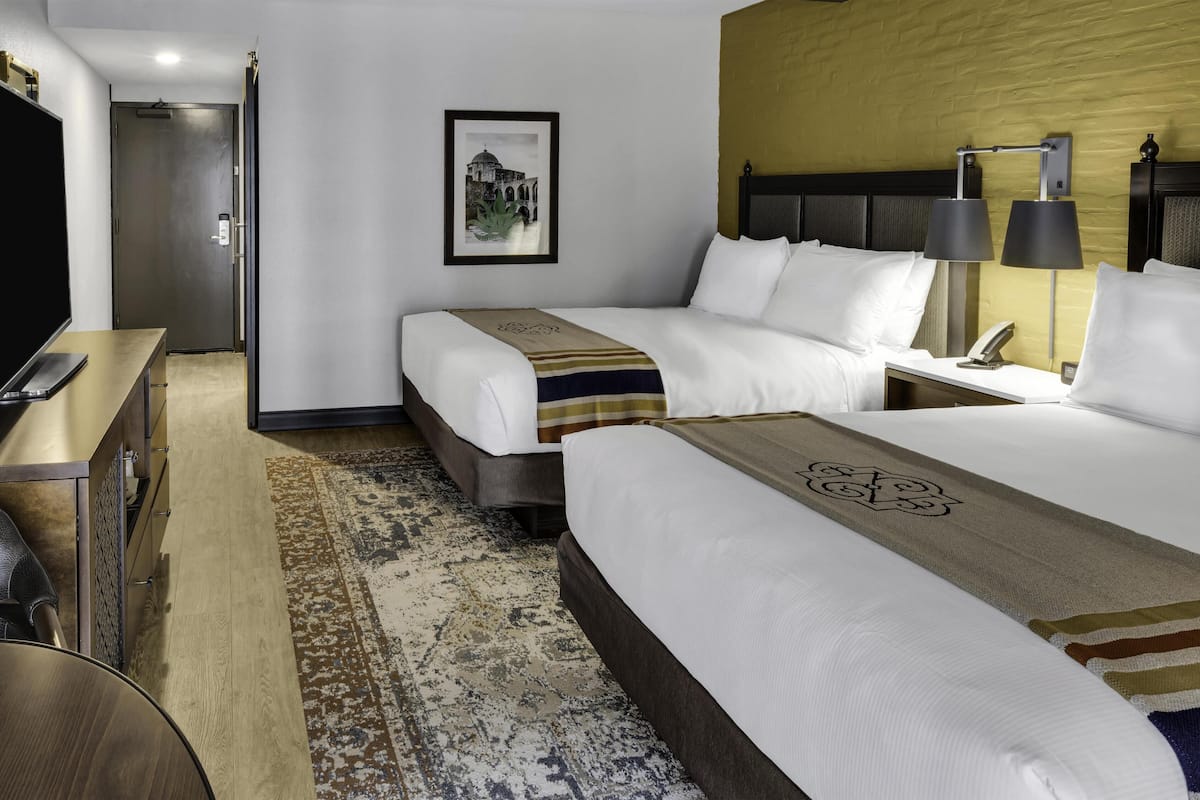 A hotel room with two beds, a desk, a TV, a framed picture, and a lamp.