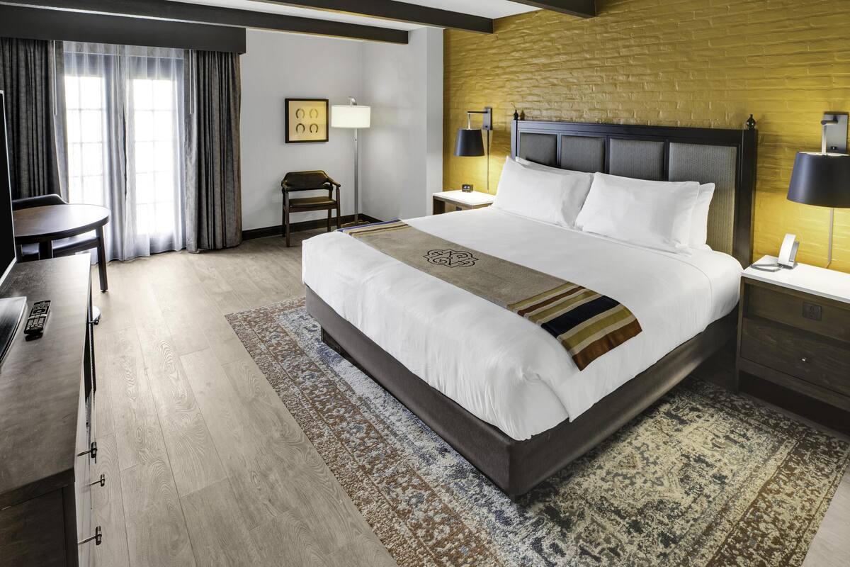 A hotel room with a large bed, a desk, a chair, a lamp, and a telephone on the nightstand.